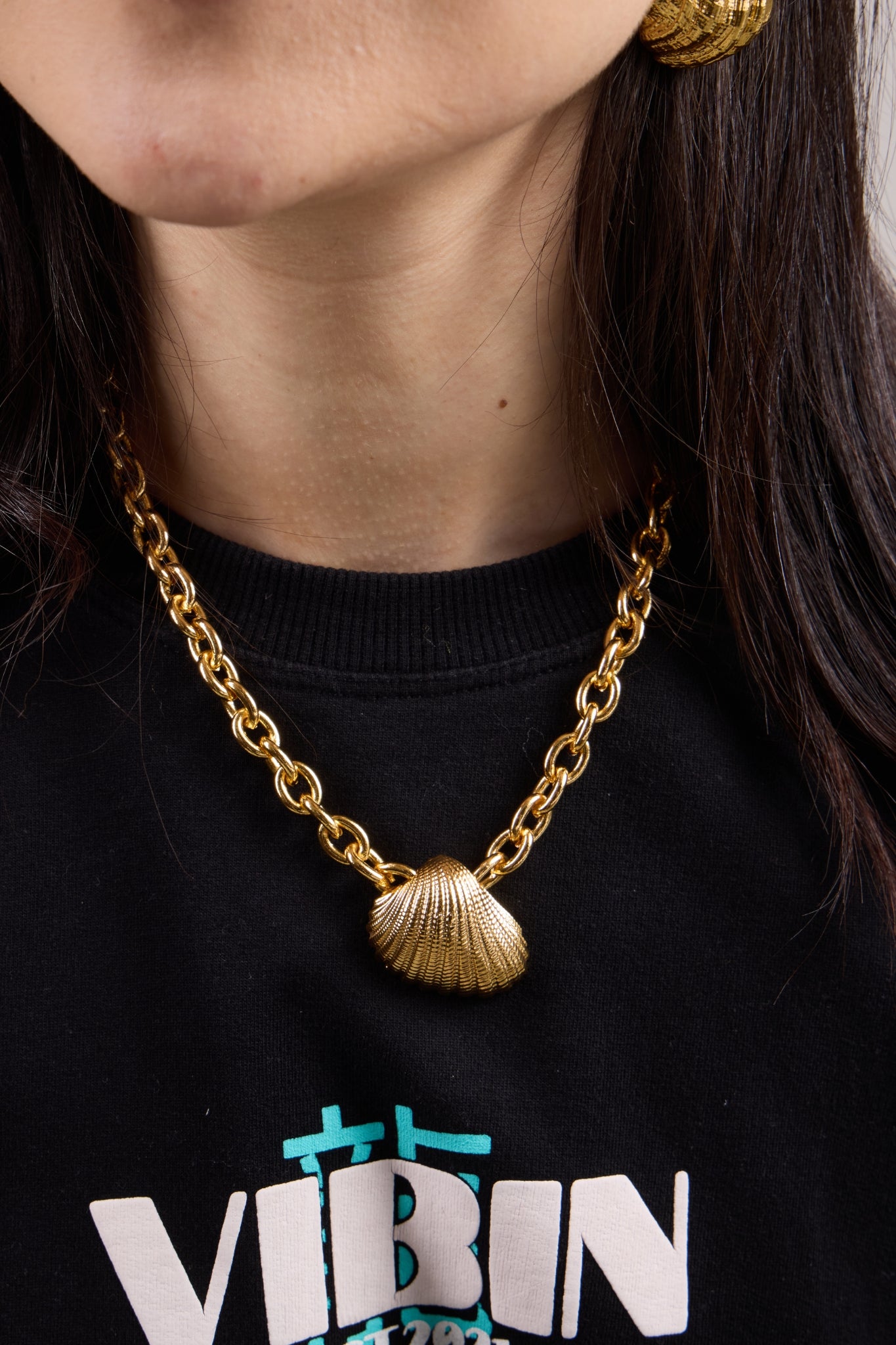 Person wearing a gold chain necklace with a shell pendant on a black shirt.