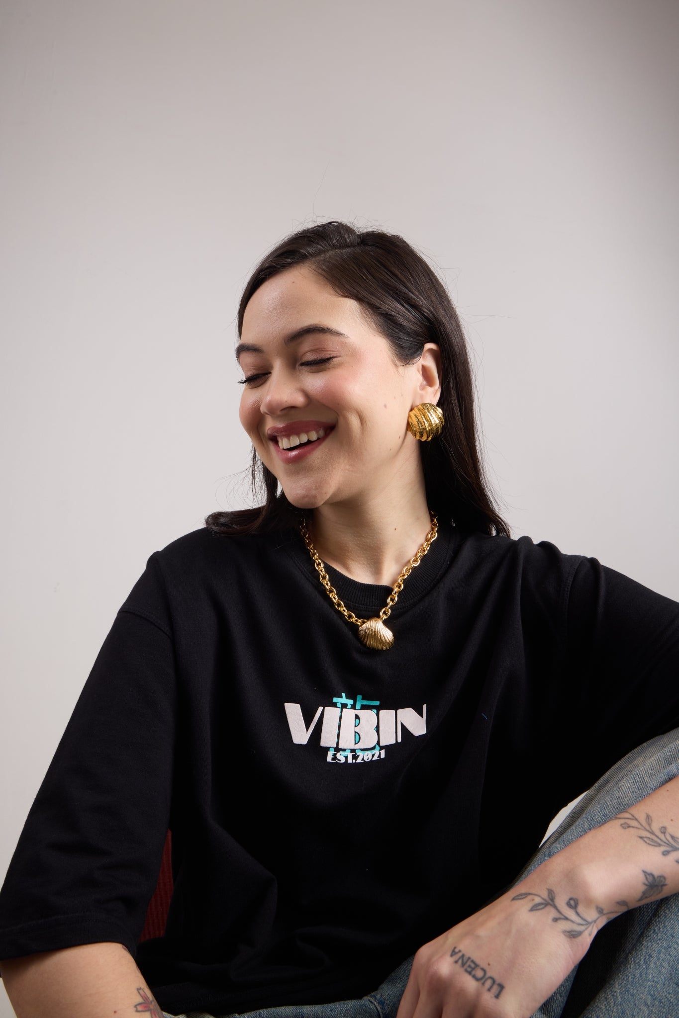 Woman wearing a gold chain necklace with a shell pendant against a plain background while sitting.
