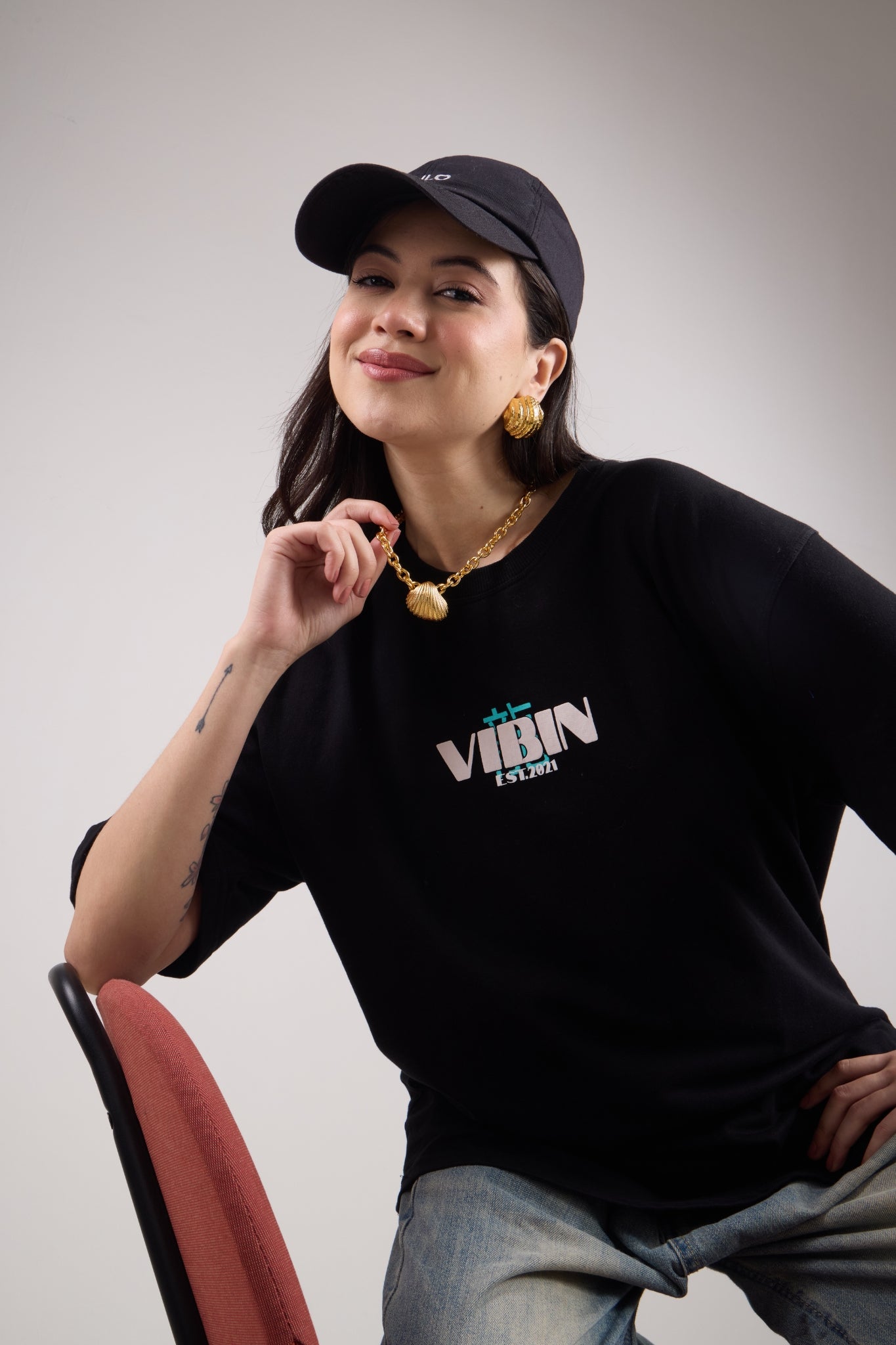 Woman wearing a gold chain necklace with a shell pendant, sitting on a chair against a plain background