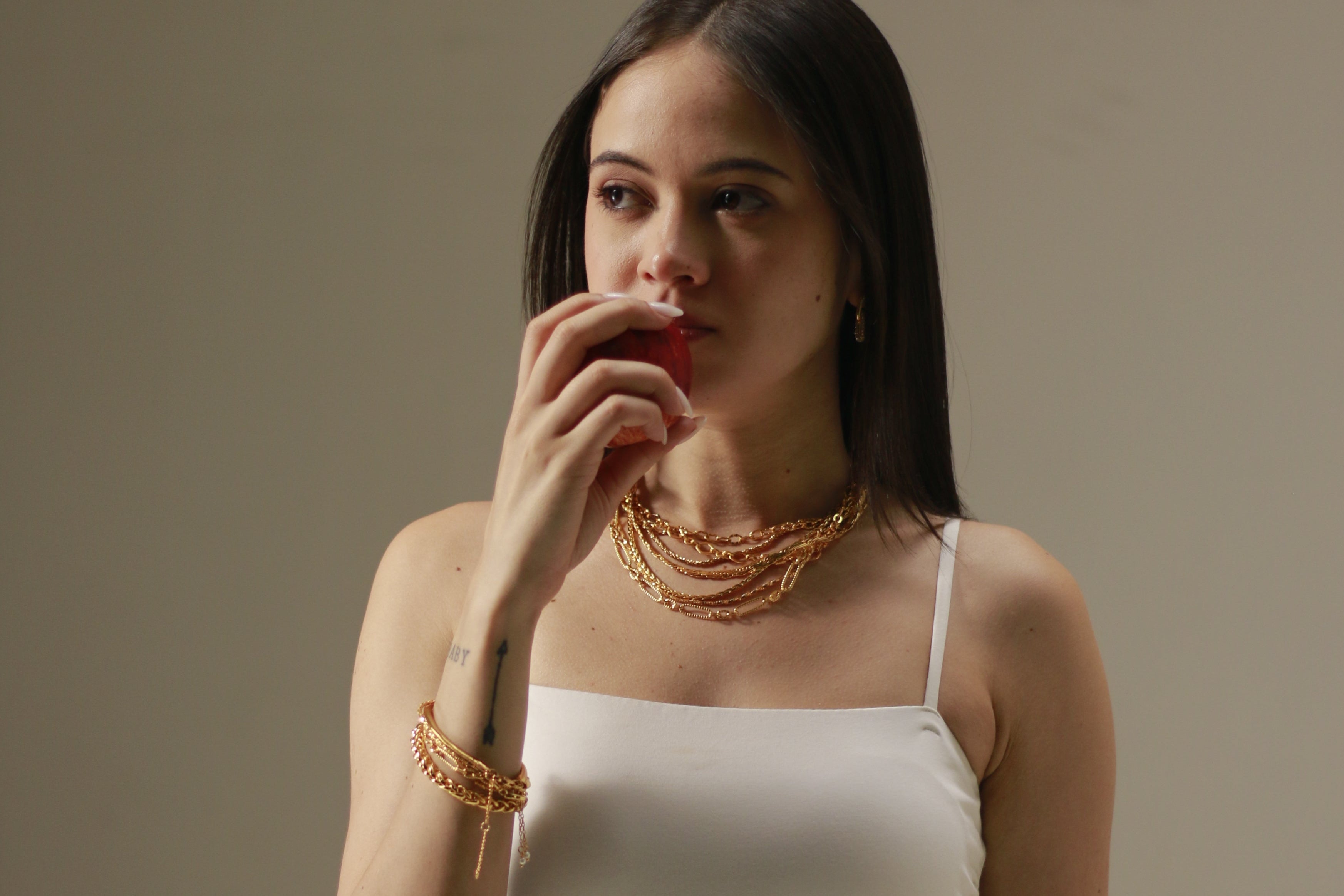 A woman with long dark hair wearing a white spaghetti-strap dress and layered gold necklaces and bracelets, holding a small red object near her mouth against a plain beige studio background.